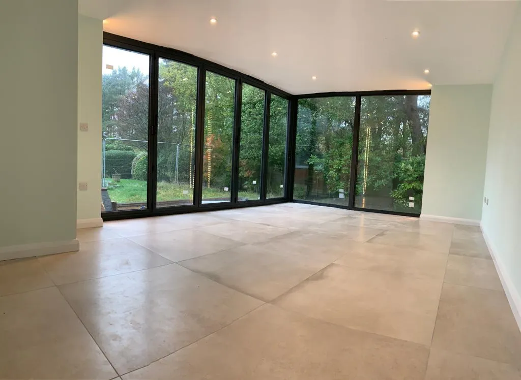 Bright and spacious room with large glass windows overlooking a green garden, featuring beige tiled flooring and light-coloured walls.