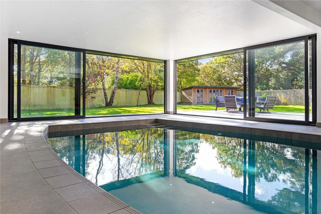 Indoor swimming pool with large glass windows, overlooking a garden with trees and a lounge area. Sunlight reflects on water.