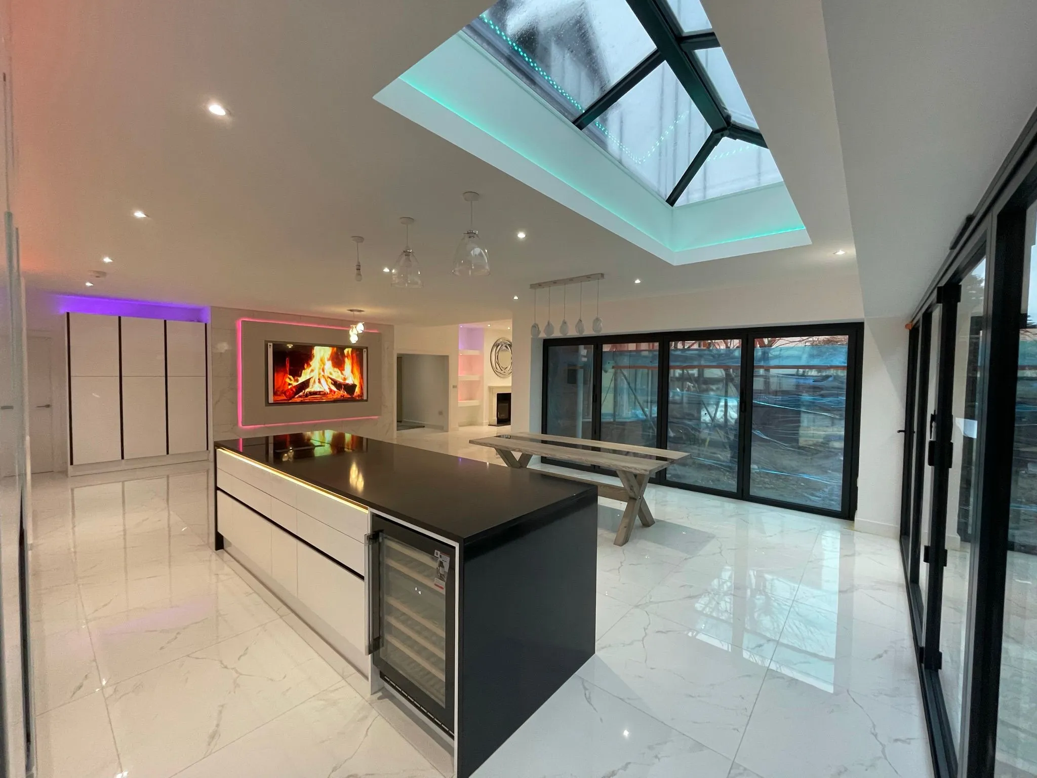 Modern kitchen with sleek white cabinets, a central island, a TV displaying a fire, and large glass windows. Ambient lighting adds color.