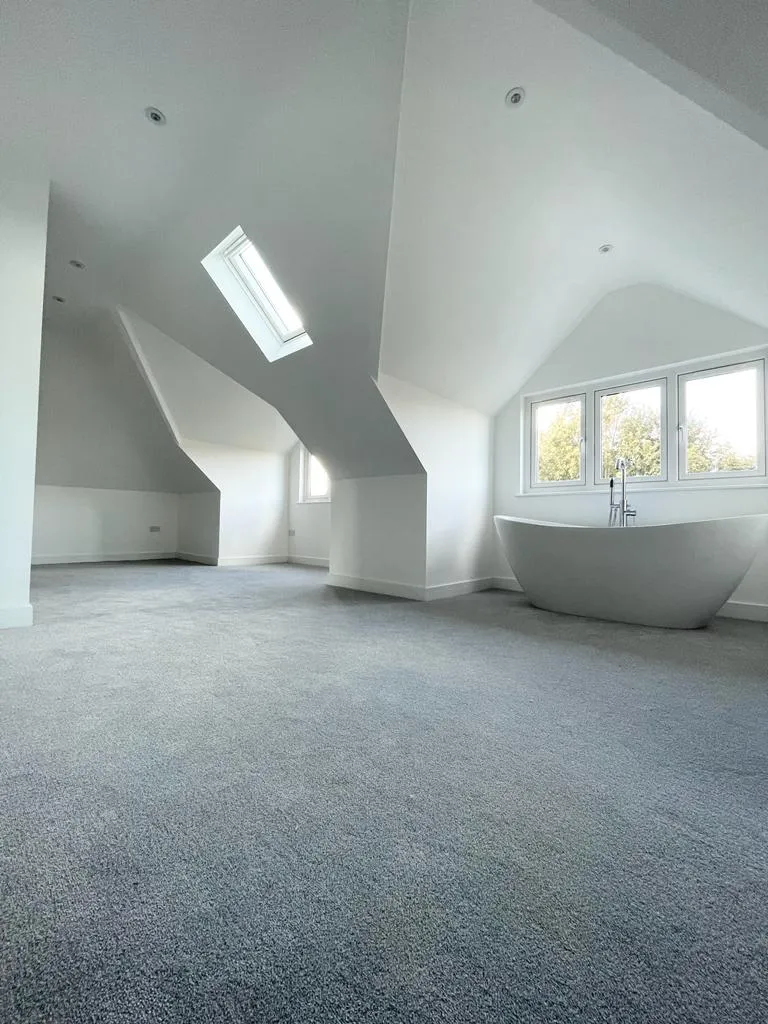 Spacious, well-lit room with sloped ceilings, skylight, large windows, and a modern freestanding bathtub on plush carpet.