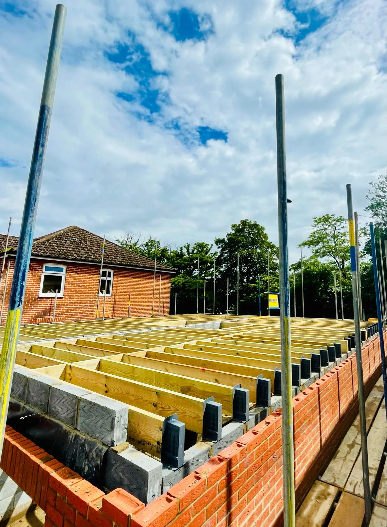 Construction site featuring wooden beams laid on a brick foundation, surrounded by scaffolding and a blue sky with clouds.